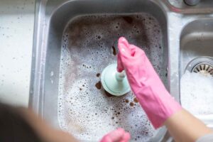 Woman using plunger in kitchen sink to unclog drain with standing water