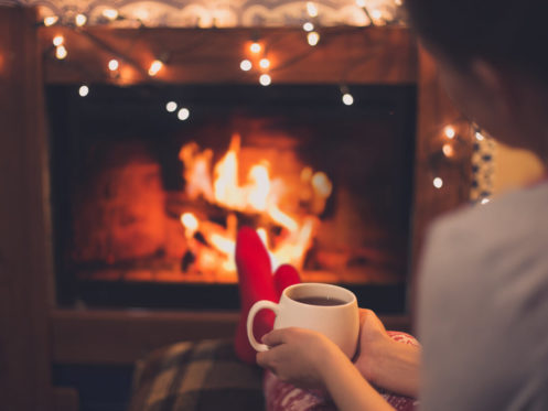 Close up cup of hot tea in woman's hands sitting near fireplace with festive Christmas lights in cozy room.